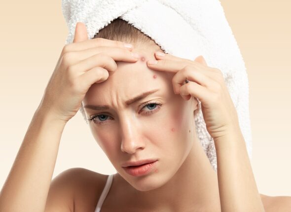 Headshot of displeased young blond woman with towel on her head, looking with painful face at the camera while squeezing pimple on her forehead. Portrait of Caucasian girl against blue wall background